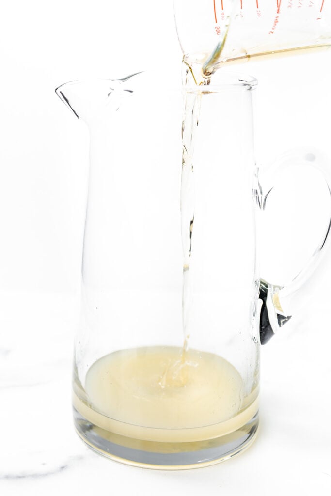 A clear liquid is being poured from a measuring cup into a glass pitcher containing a small amount of yellowish liquid.