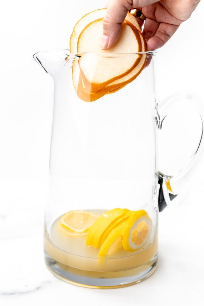 A hand places thin pear slices into a glass pitcher containing lemon slices and a small amount of juice.