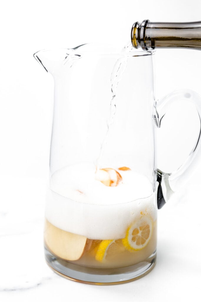 A clear glass pitcher with sliced apples and lemons is being filled with a fizzy beverage from a bottle.