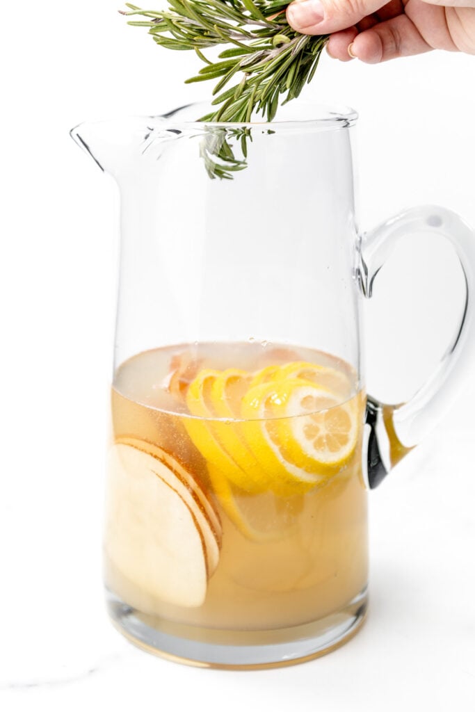 A glass pitcher filled with sliced lemons and apples in liquid, with a hand holding rosemary sprigs above the pitcher.