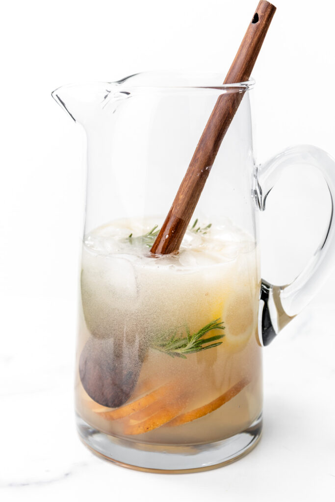 A glass pitcher filled with ice, citrus slices, herbal sprigs, and a light-colored beverage, with a wooden stirring stick inside.