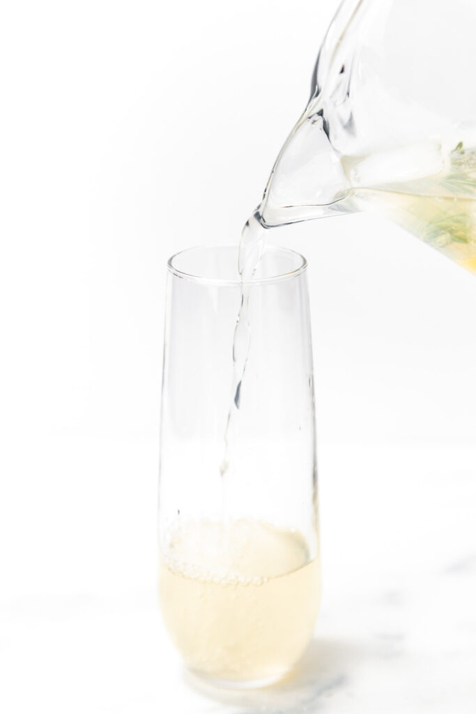 A clear liquid is being poured from a glass pitcher into a tall, narrow glass with a light-colored drink inside.