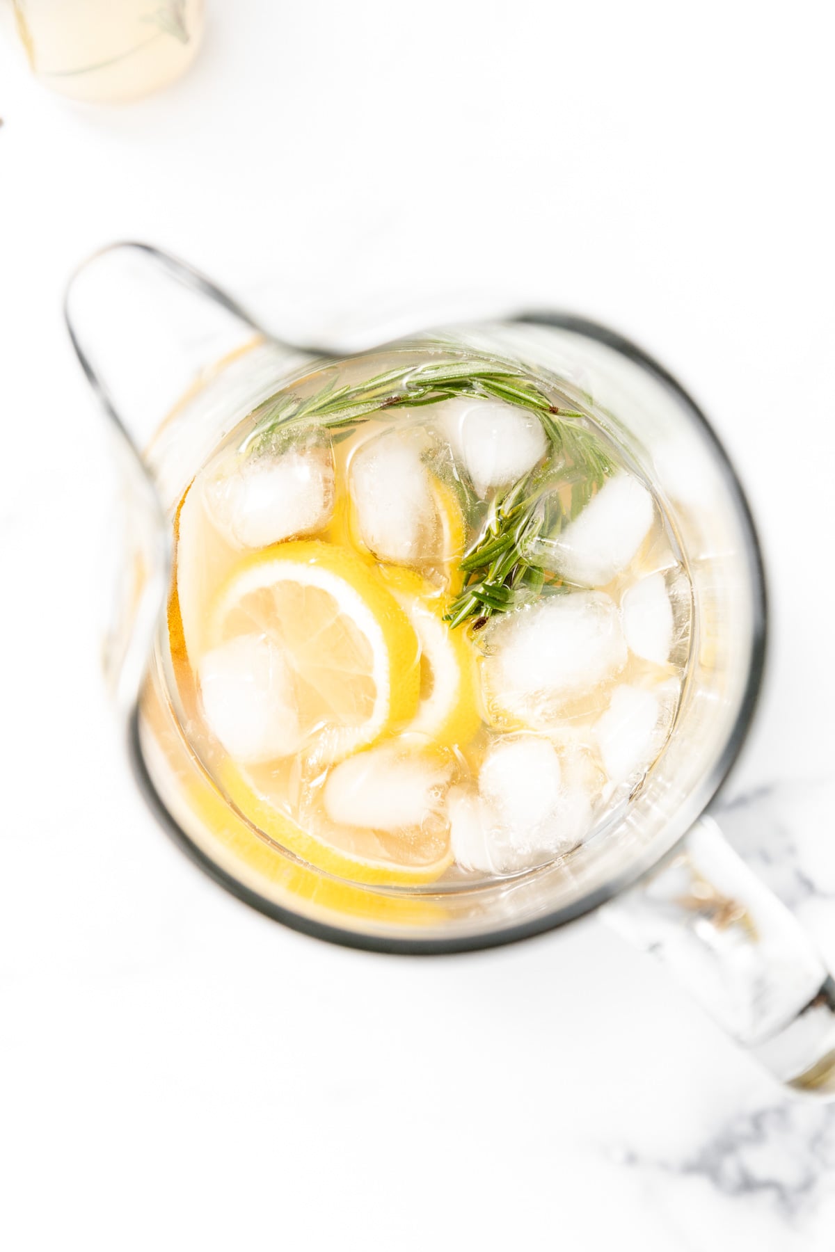 A glass pitcher filled with ice, lemon slices, rosemary sprigs, and a light-colored beverage, viewed from above on a white surface.
