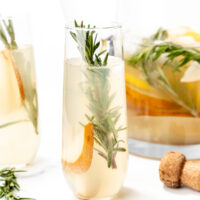A glass of light-colored cocktail garnished with a sprig of rosemary and a pear slice, with a cork and a pitcher of similar drinks in the background.