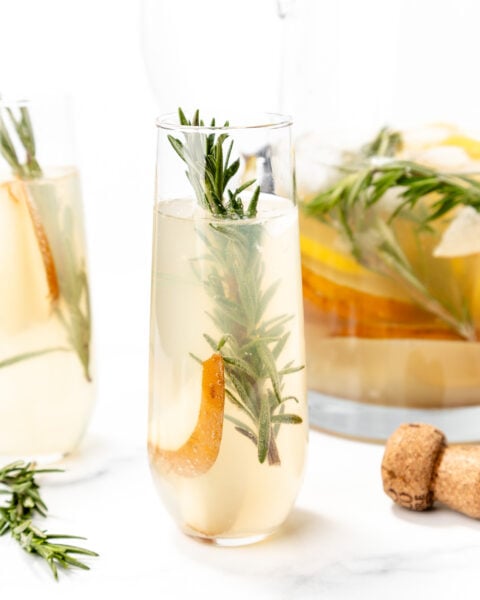 A glass of light-colored cocktail garnished with a sprig of rosemary and a pear slice, with a cork and a pitcher of similar drinks in the background.