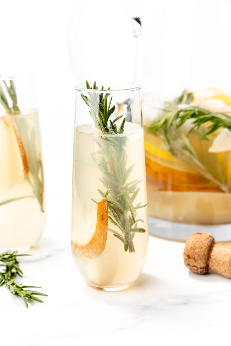 A glass of light-colored cocktail garnished with a sprig of rosemary and a pear slice, with a cork and a pitcher of similar drinks in the background.