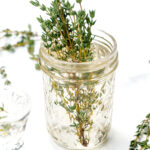 A small glass jar filled with water holds fresh thyme sprigs, with more thyme scattered on a white surface nearby.