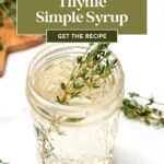 A glass jar filled with clear thyme simple syrup, garnished with fresh thyme sprigs, sits on a white surface. Text overlay reads: "Thyme Simple Syrup - Get the Recipe.