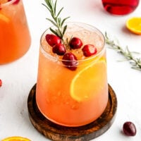 A glass of iced orange-red drink garnished with cranberries, an orange slice, and a sprig of rosemary, set on a wooden coaster with fruit and rosemary nearby.