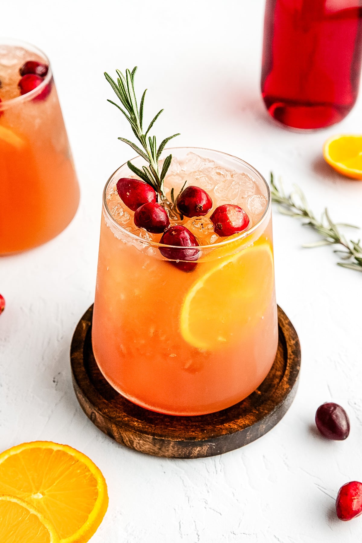 A glass of iced orange-red drink garnished with cranberries, an orange slice, and a sprig of rosemary, set on a wooden coaster with fruit and rosemary nearby.