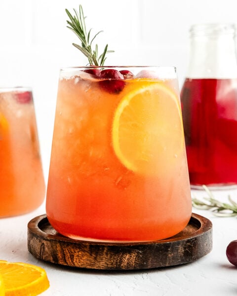 A glass of iced orange and red fruit drink garnished with a slice of orange, cranberries, and a sprig of rosemary, on a wooden coaster.