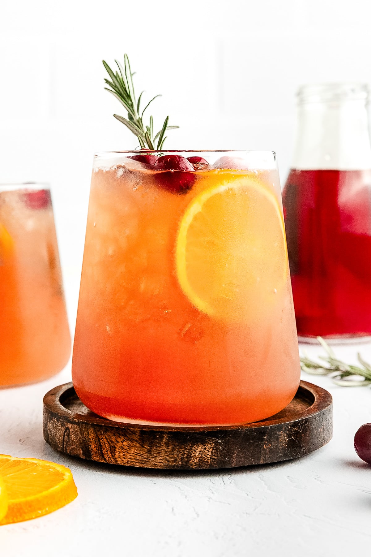 A glass of iced orange and red fruit drink garnished with a slice of orange, cranberries, and a sprig of rosemary, on a wooden coaster.