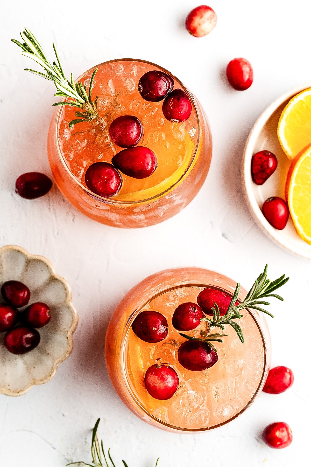 Two glasses of iced orange-colored drink garnished with cranberries and rosemary, surrounded by loose cranberries and orange slices on a white surface.