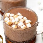 A glass mug of hot chocolate topped with mini marshmallows, placed on a wooden coaster with scattered marshmallows nearby.