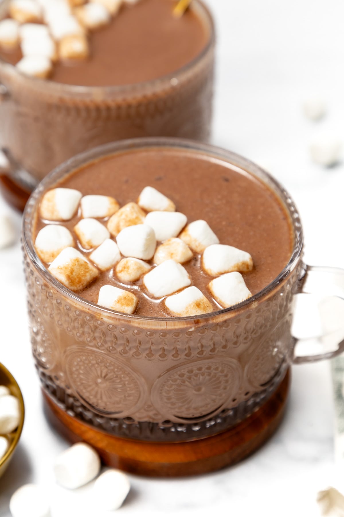 A glass mug of hot chocolate topped with mini marshmallows, placed on a wooden coaster with scattered marshmallows nearby.