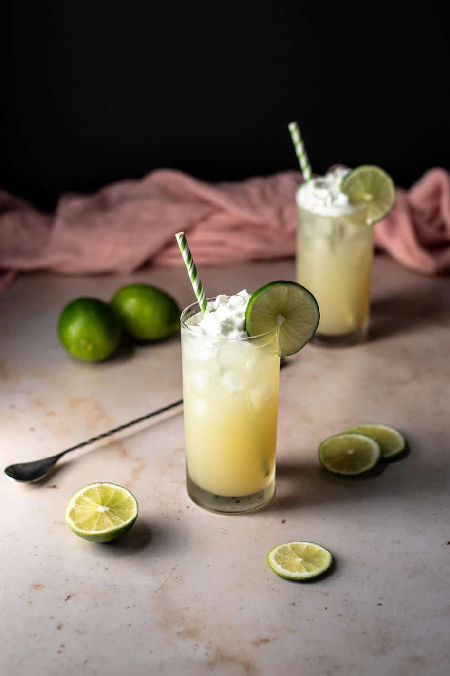 Two tall glasses filled with refreshing green cocktails, ice, whipped cream, and lime slices, each with striped straws. Whole and sliced limes are scattered on the surface nearby.