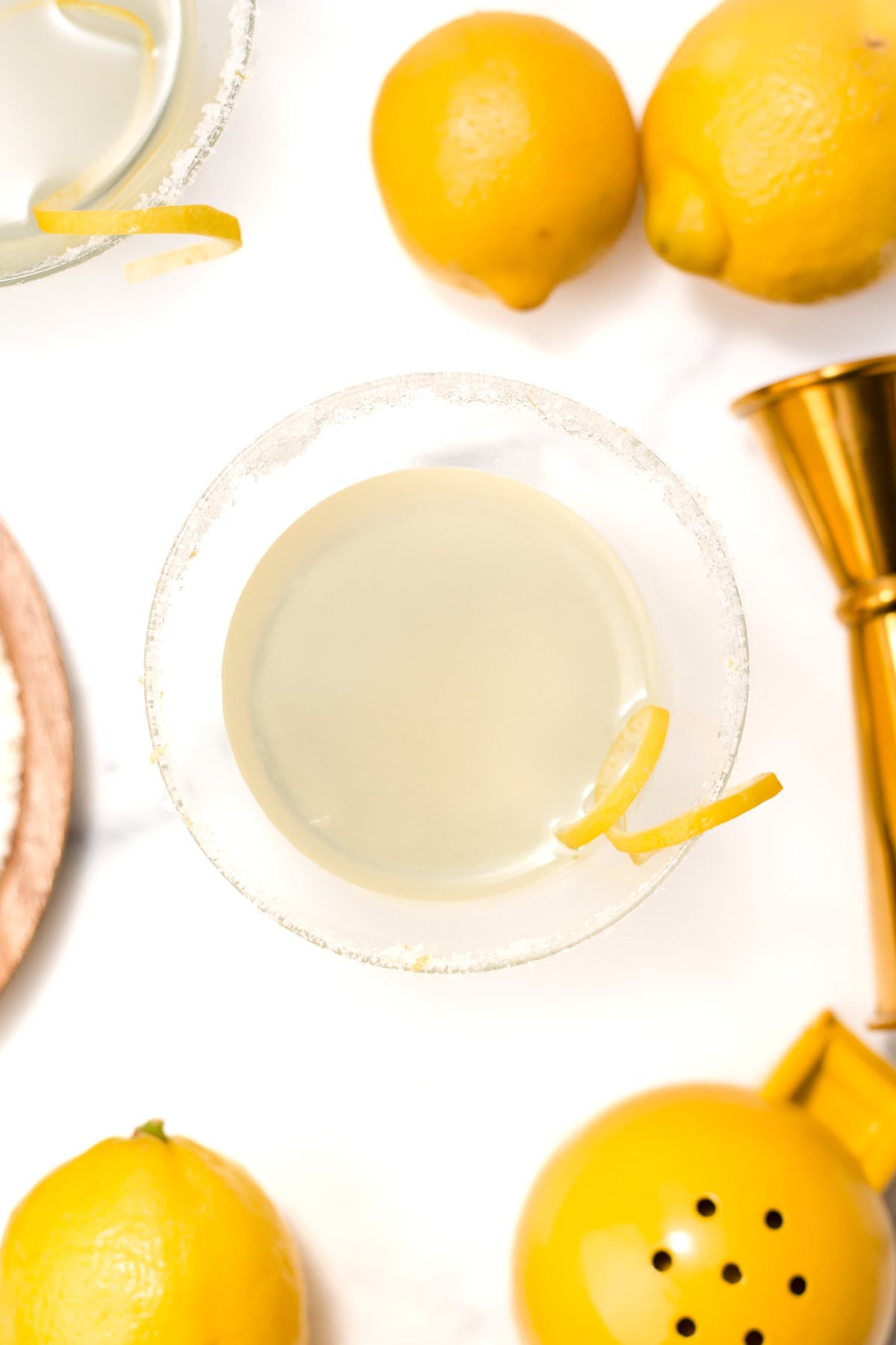 A cocktail in a sugar-rimmed glass with a lemon twist garnish, surrounded by whole lemons, a jigger, and a lemon squeezer on a white surface.