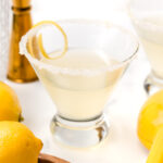 A lemon cocktail in a glass with a sugared rim and a lemon twist, surrounded by fresh lemons and cocktail tools on a white surface.