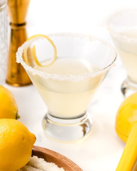 A lemon cocktail in a glass with a sugared rim and a lemon twist, surrounded by fresh lemons and cocktail tools on a white surface.