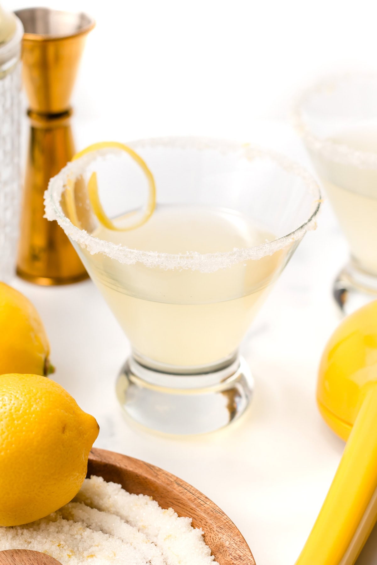 A lemon cocktail in a glass with a sugared rim and a lemon twist, surrounded by fresh lemons and cocktail tools on a white surface.