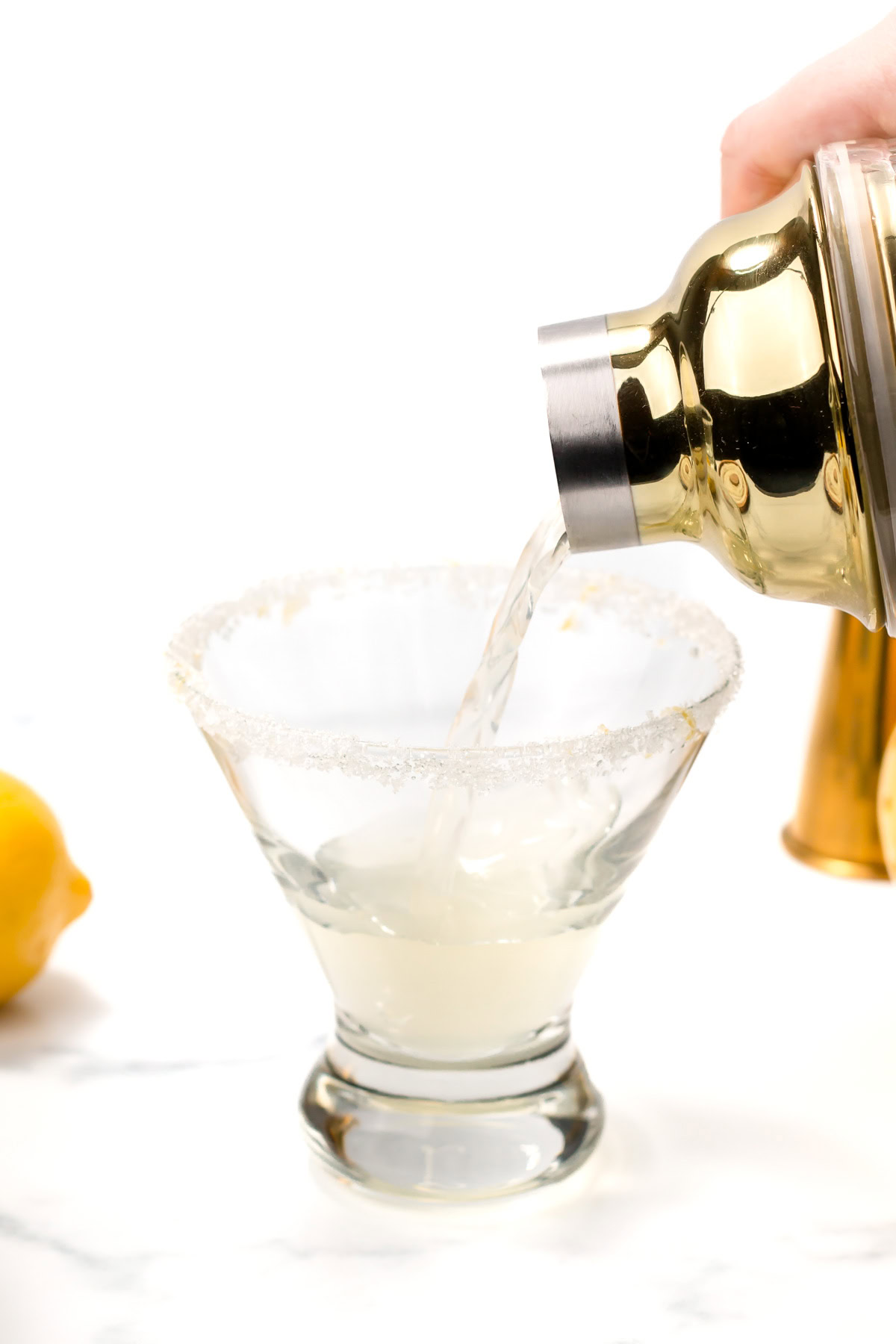 A cocktail shaker pours a clear liquid into a sugar-rimmed glass, with a lemon and jigger in the background on a white surface.