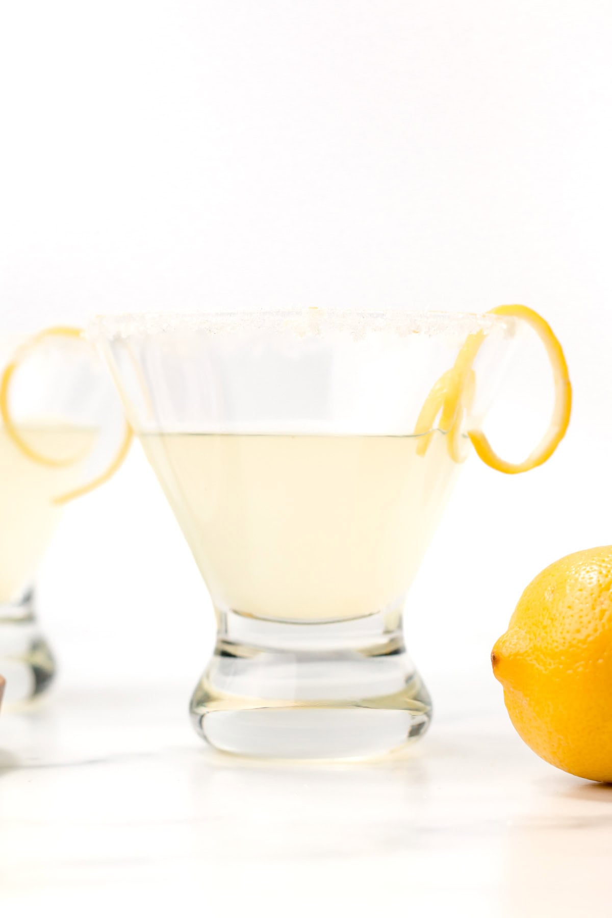 A martini glass filled with a pale yellow drink, garnished with a sugared rim and a lemon twist, with a whole lemon nearby.