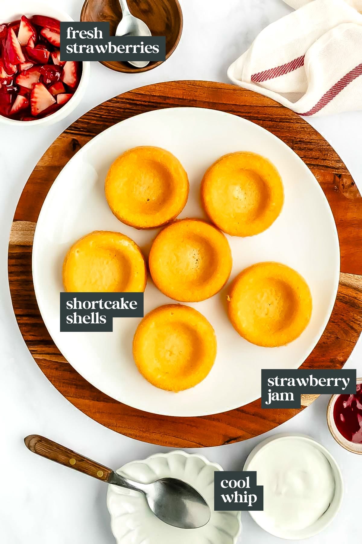 A plate of six mini strawberry shortcakes sits on a wooden tray, surrounded by bowls of fresh strawberries, strawberry jam, and cool whip.