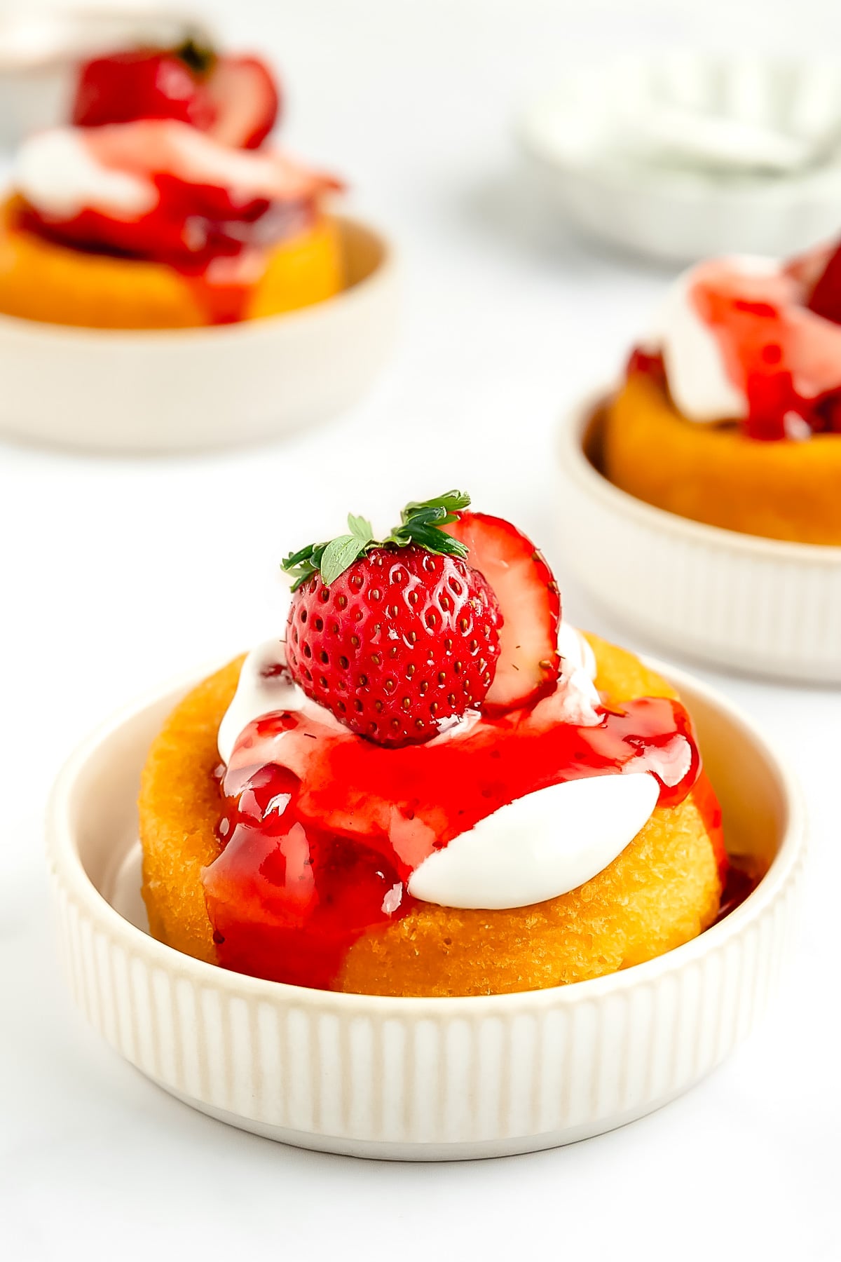 A dessert of shortcake topped with whipped cream, strawberry sauce, and a whole strawberry, served in a small white dish.