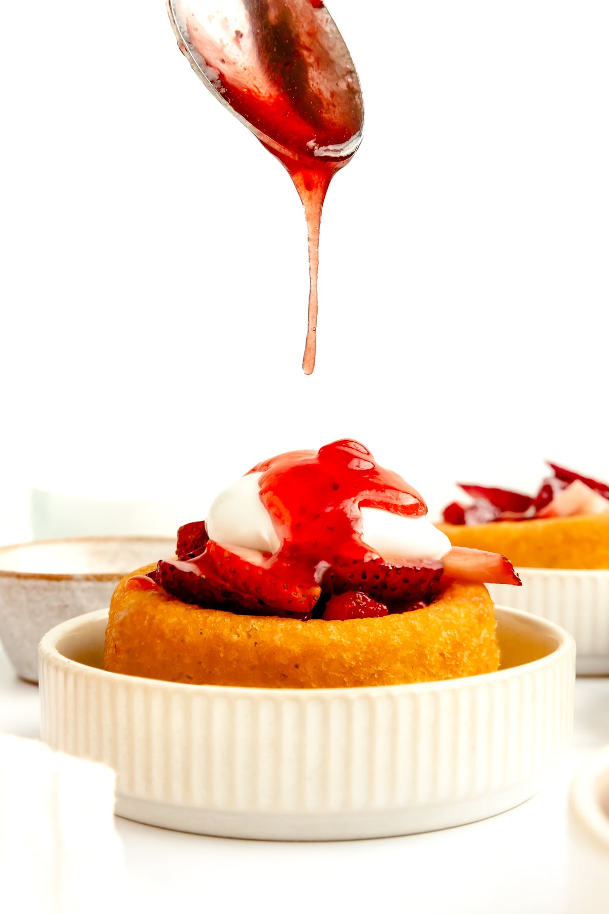 A dessert topped with strawberries and cream in a white bowl, with strawberry sauce being drizzled from a spoon above.