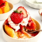 A small round sponge cake topped with whipped cream, strawberry sauce, and a whole strawberry, served in a beige bowl with a fork.