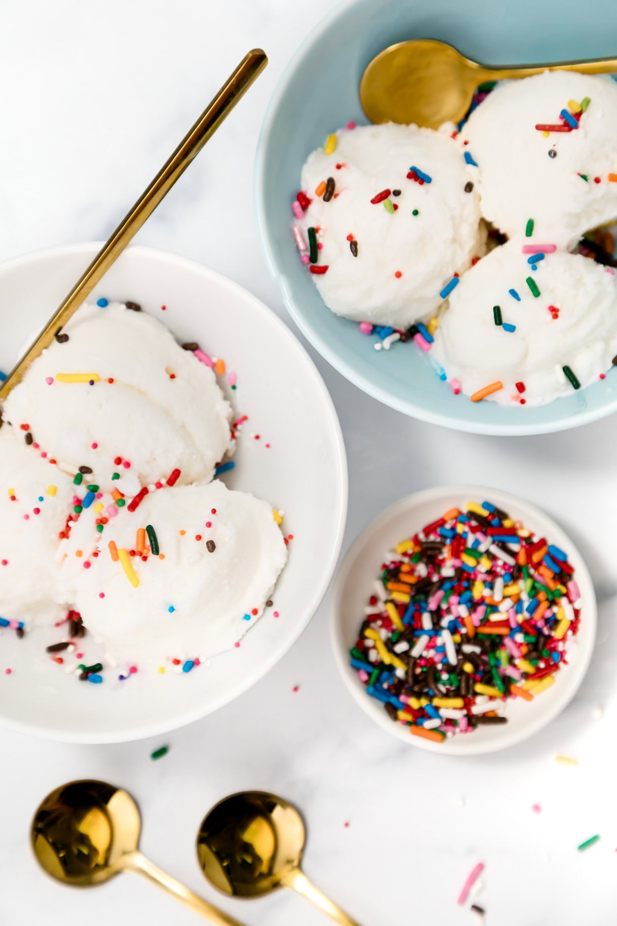 Two bowls of vanilla ice cream with rainbow sprinkles and gold spoons, next to a small dish of extra sprinkles on a white surface.