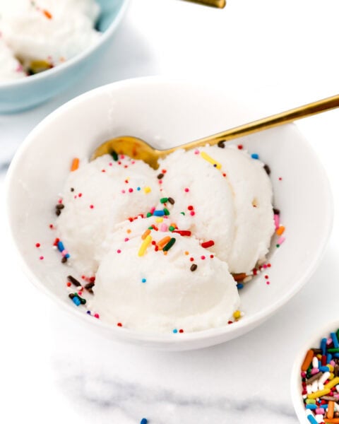 A white bowl with three scoops of vanilla ice cream topped with colorful sprinkles and a gold spoon, with another bowl of ice cream and a dish of sprinkles nearby.