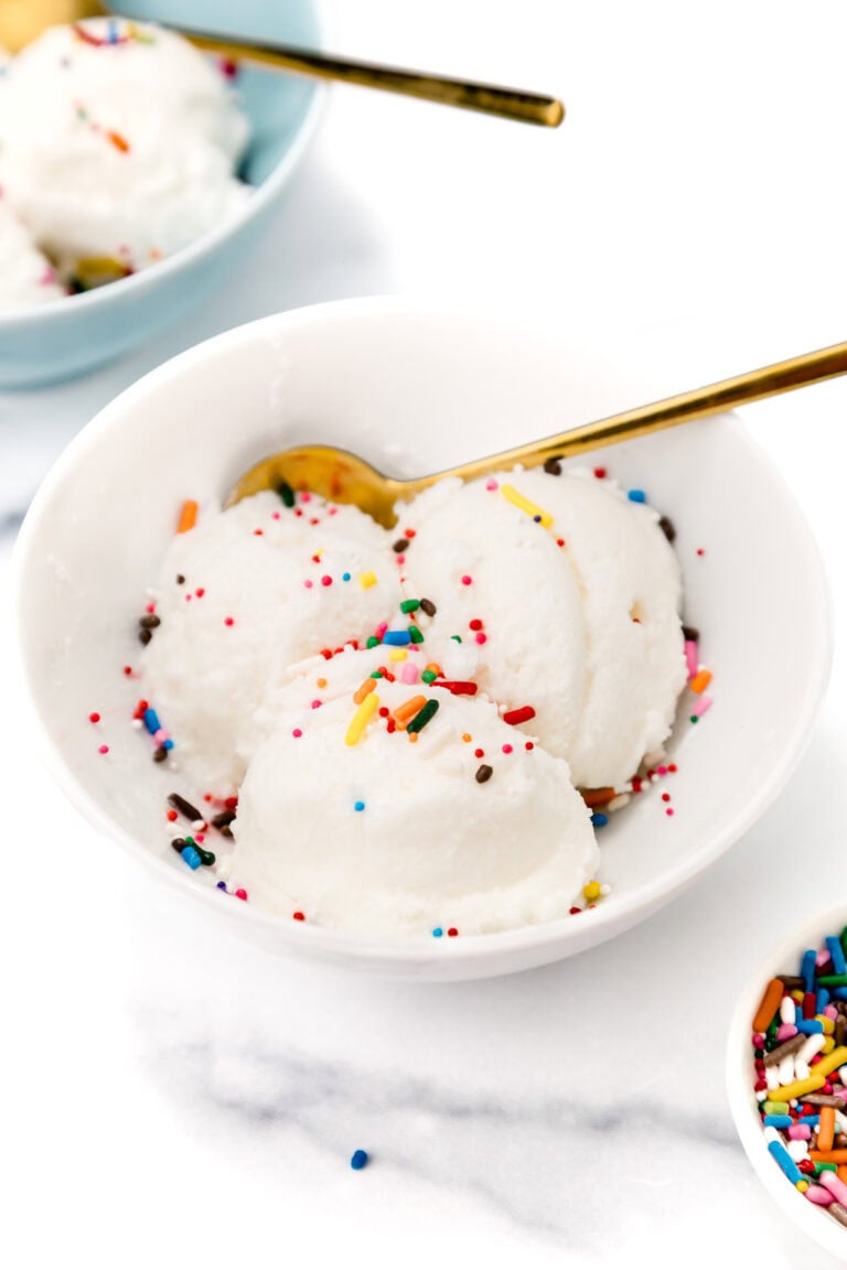 A white bowl with three scoops of vanilla ice cream topped with colorful sprinkles and a gold spoon, with another bowl of ice cream and a dish of sprinkles nearby.