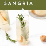 A glass of sparkling sangria garnished with rosemary and fruit sits on a white surface next to a cork, with text promoting a recipe.