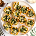 A plate of spinach and cheese-filled wonton cups is arranged on a white surface, surrounded by bowls of grated cheese and chili flakes.