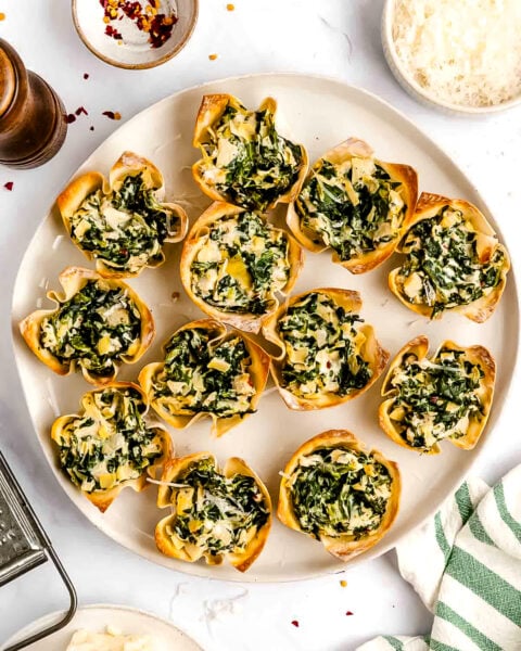 A plate of spinach and cheese-filled wonton cups is arranged on a white surface, surrounded by bowls of grated cheese and chili flakes.