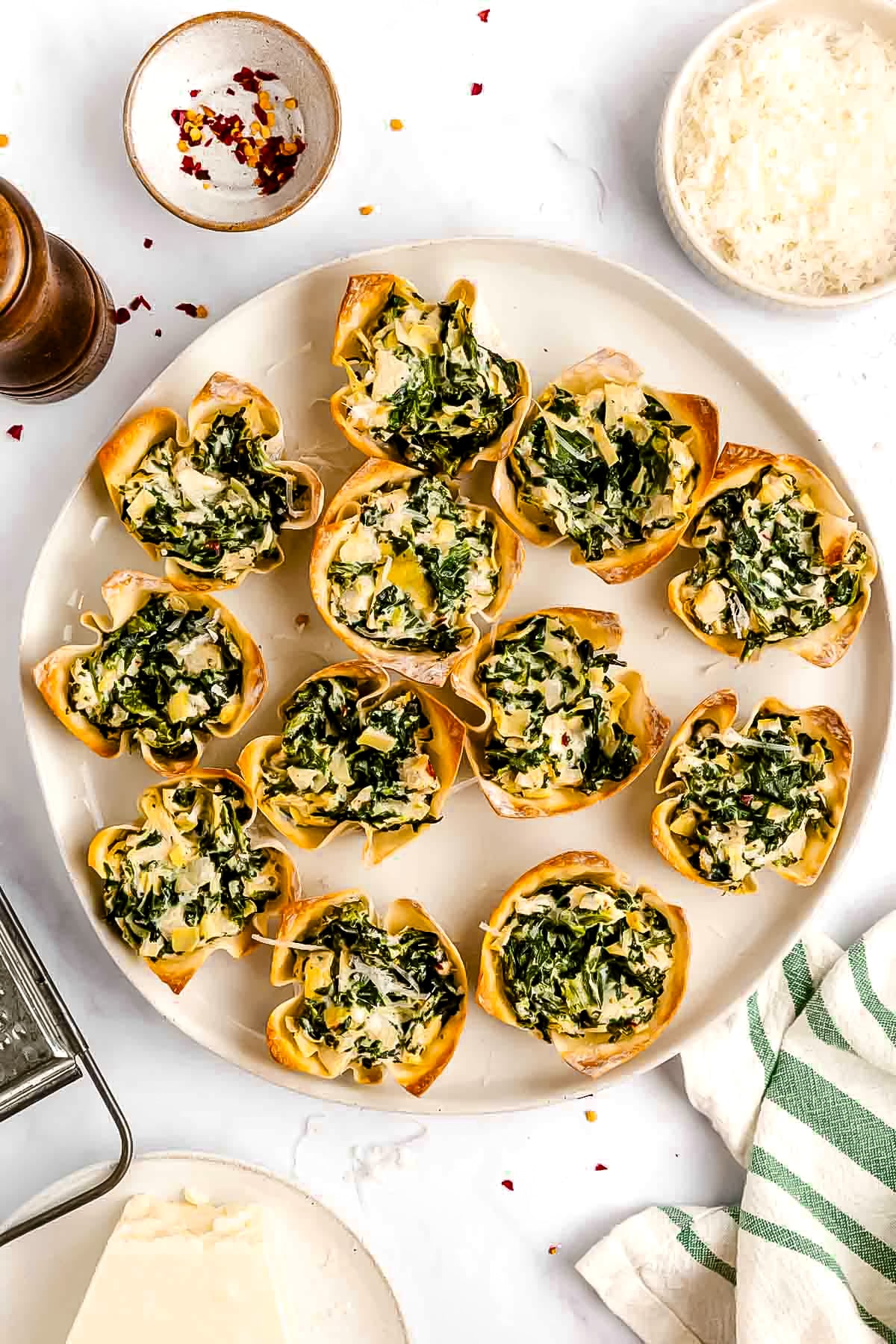 A plate of spinach and cheese-filled wonton cups is arranged on a white surface, surrounded by bowls of grated cheese and chili flakes.