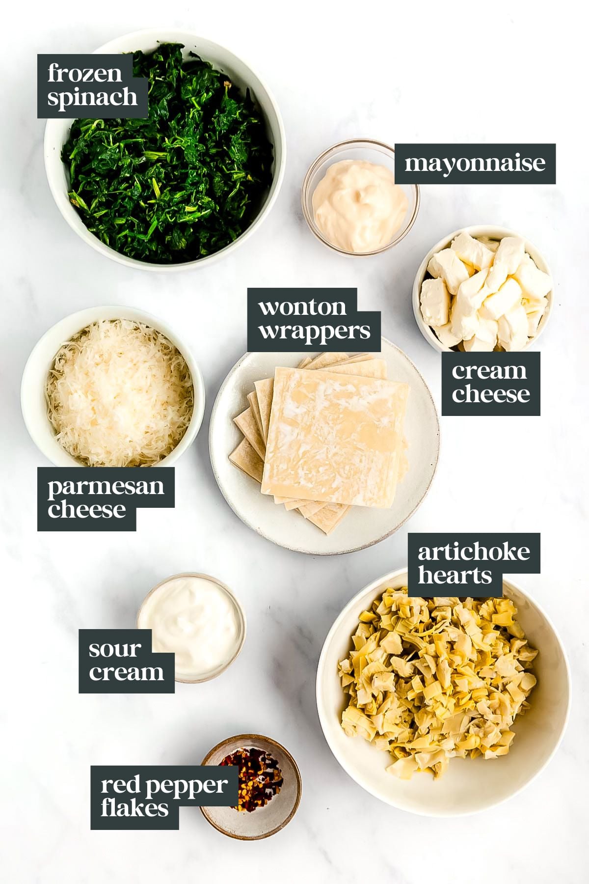 Bowls of frozen spinach, mayonnaise, cream cheese, parmesan cheese, sour cream, artichoke hearts, red pepper flakes, and wonton wrappers are ready to create delicious spinach artichoke wonton cups on a white surface.