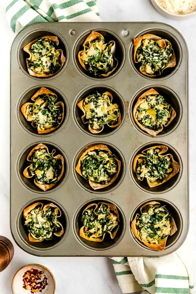 A muffin tin holds twelve spinach and cheese-filled phyllo cups, freshly baked, with a green and white striped towel nearby.