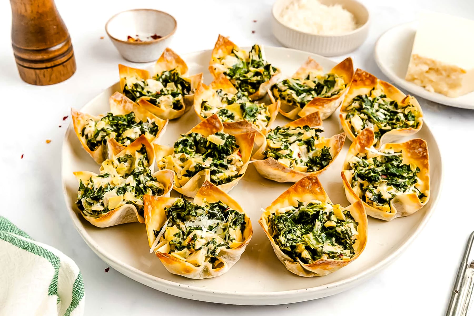 A plate of spinach and cheese mini quiches in crispy wonton cups, displayed on a white surface with bowls of grated cheese and seasoning nearby.