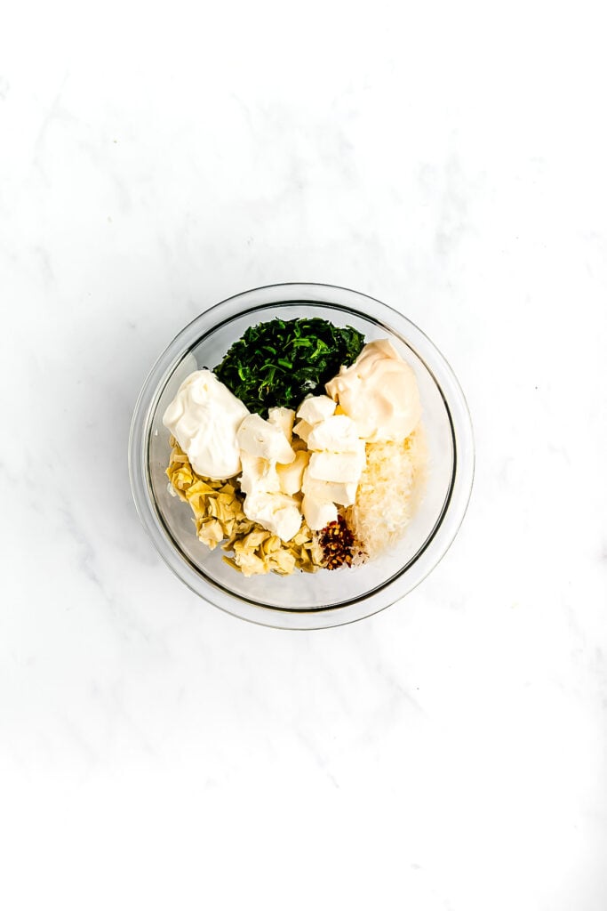 A glass bowl containing mayonnaise, chopped spinach, artichoke hearts, cream cheese, grated cheese, and minced garlic on a white surface.