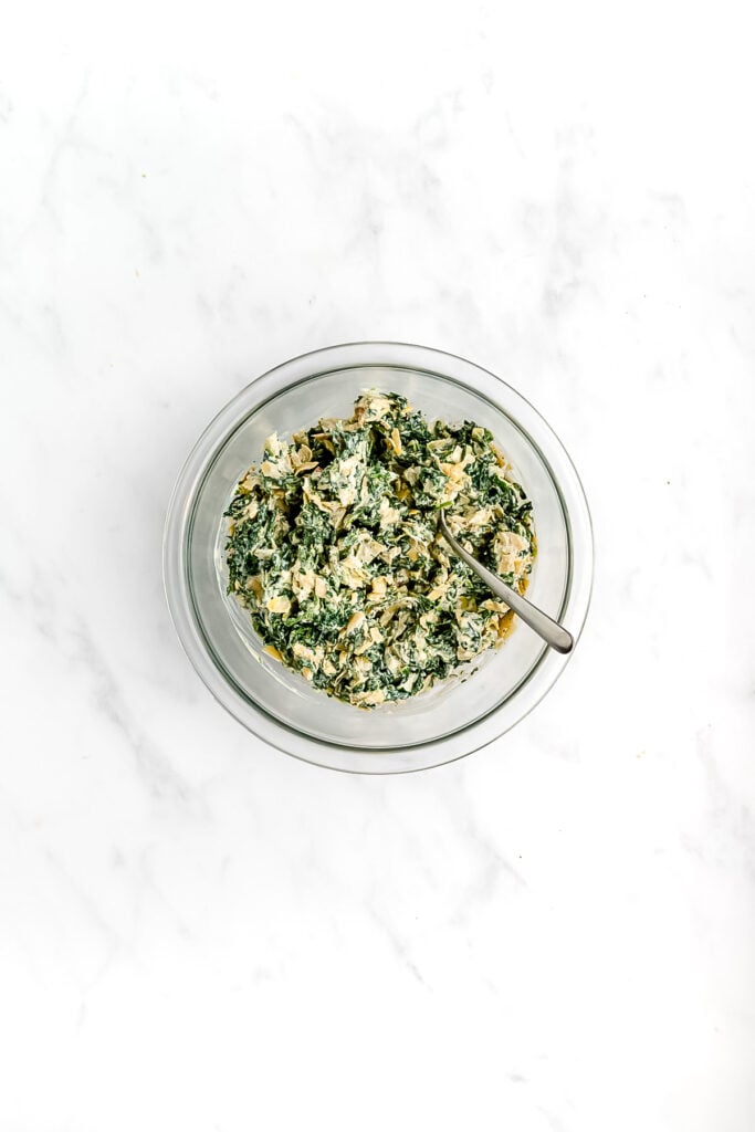 A glass bowl filled with creamy spinach dip, mixed with pieces of spinach and a spoon inside, on a white marble surface.