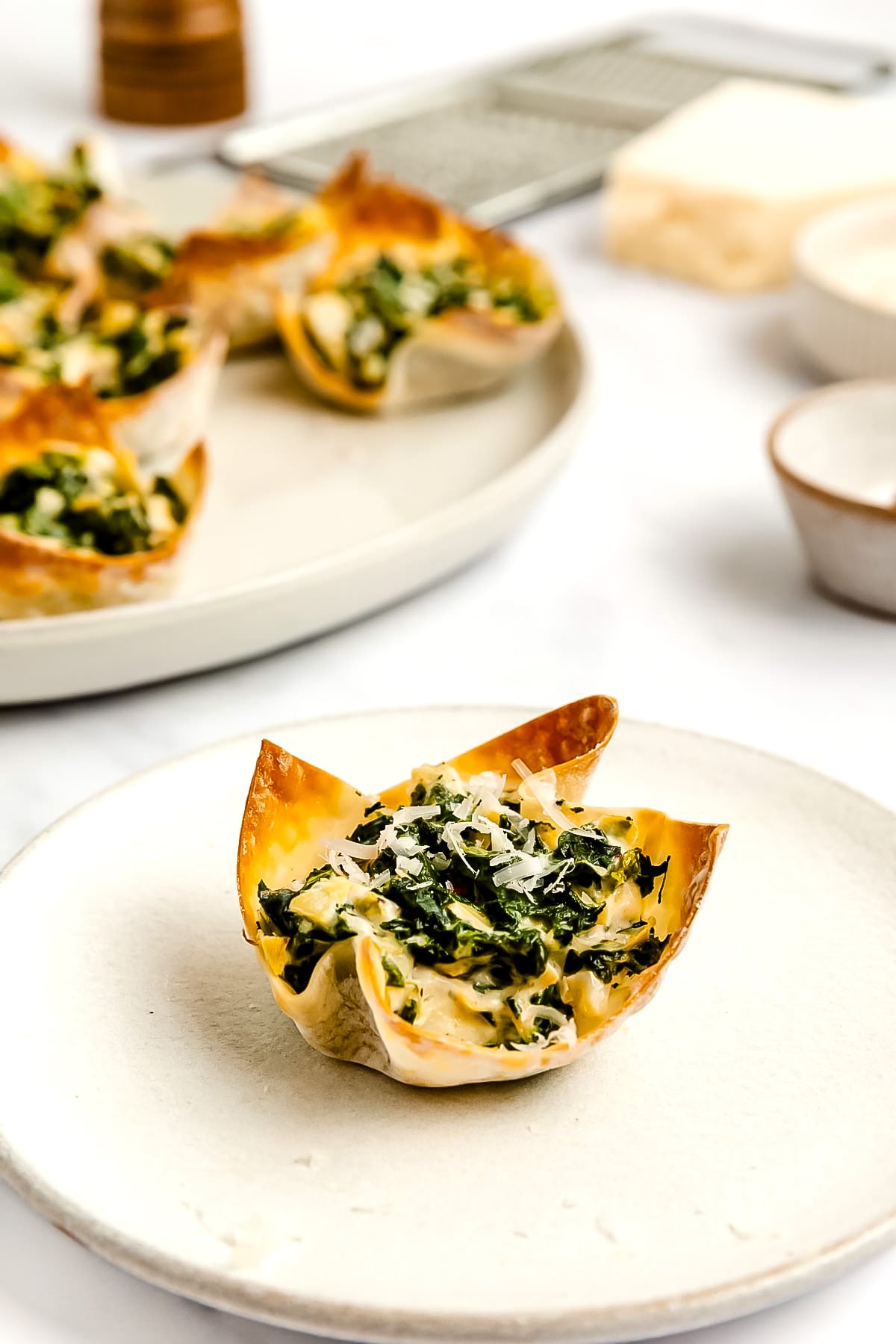 A single spinach and cheese wonton cup on a white plate, with a plate of several more wonton cups and ingredients in the background.