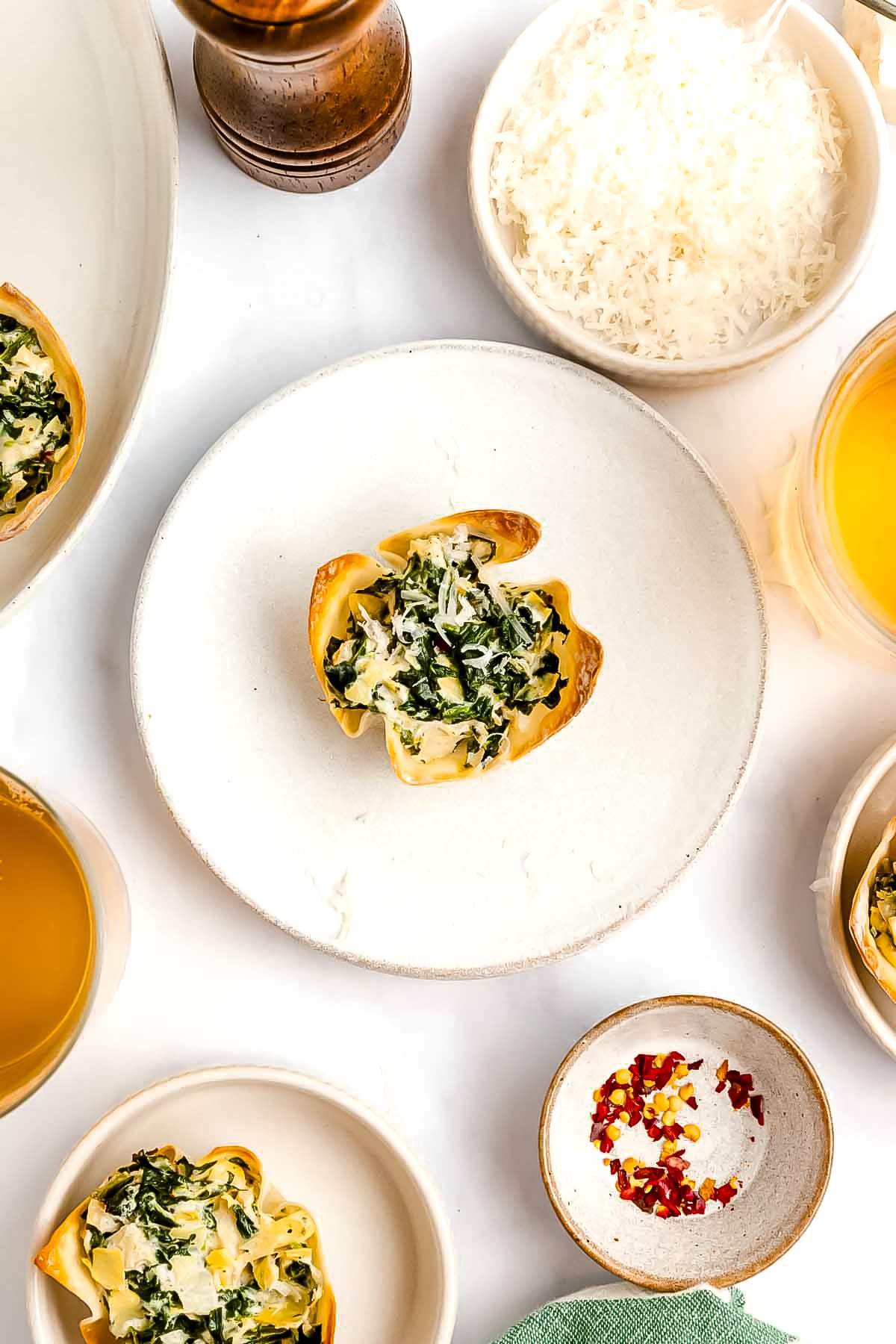 A plate with a spinach and cheese stuffed pasta shell, surrounded by bowls of shredded cheese, broth, red pepper flakes, and other ingredients on a white surface.