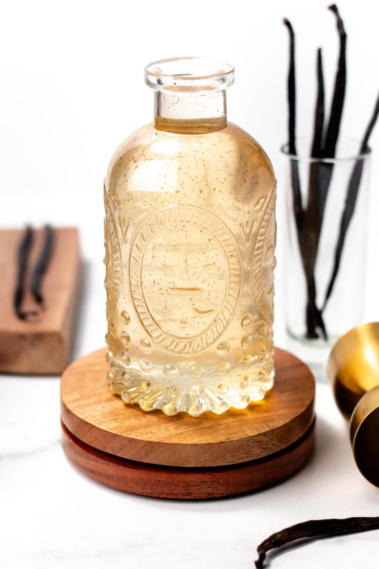 A clear glass bottle of vanilla extract with visible seeds sits on a wooden coaster, with vanilla beans and a glass cup in the background.