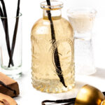 A glass bottle of vanilla extract with a vanilla bean inside, surrounded by vanilla pods, a glass, a gold jigger, and wooden utensils on a white surface.