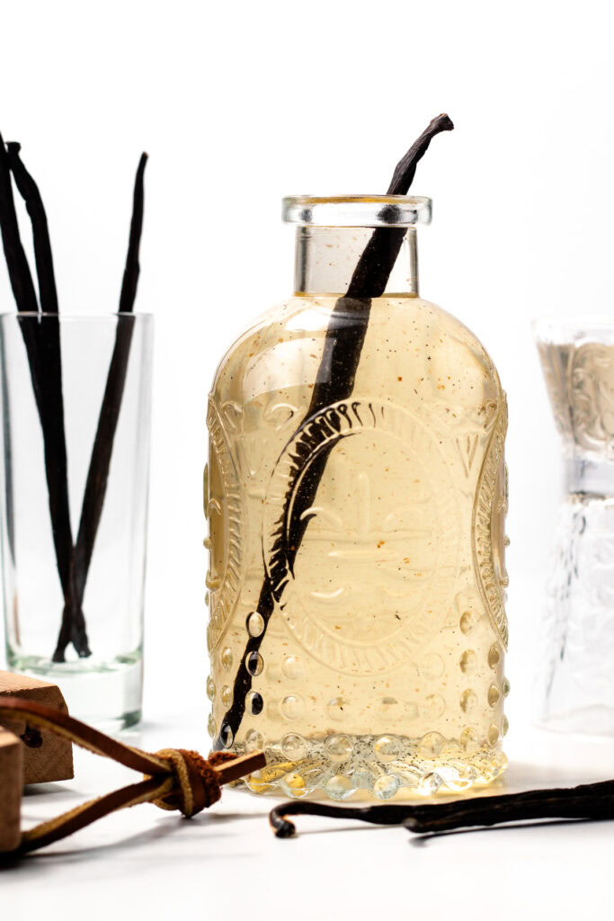 A glass bottle filled with vanilla extract, containing a vanilla bean pod, is placed in front of a glass and other vanilla pods.