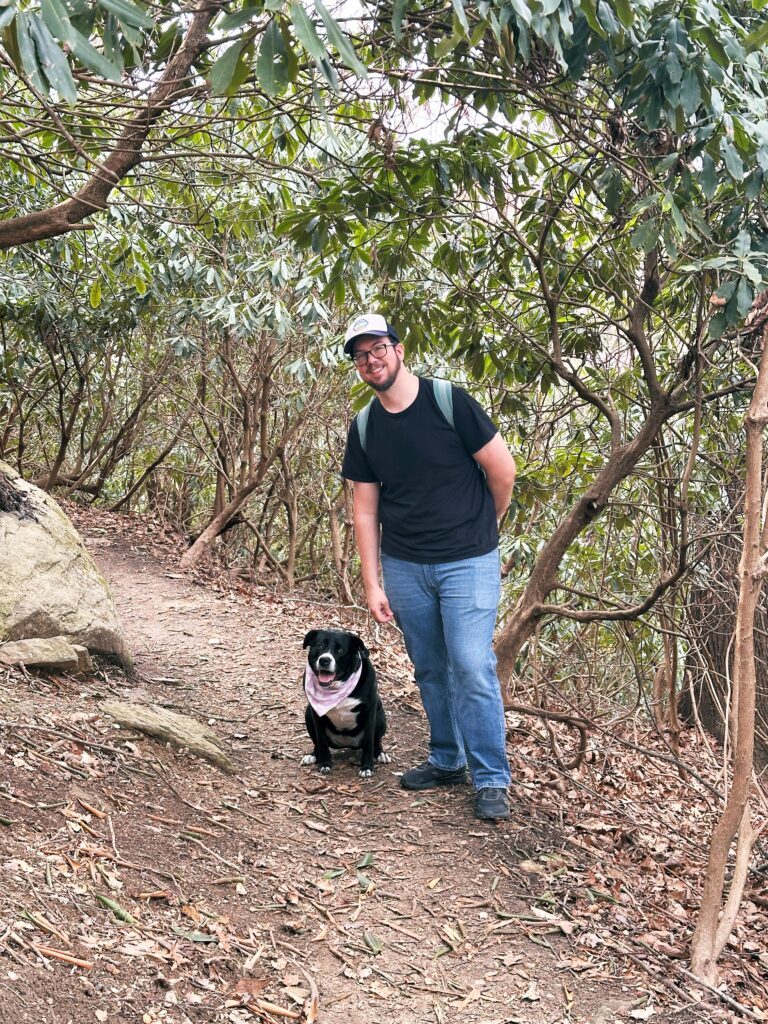 A man wearing a cap and backpack stands on a forest trail next to a black dog with a pink bandana, surrounded by trees and foliage&mdash;a serene scene reminiscent of Susannah Brinkley Henry&rsquo;s outdoor adventures.