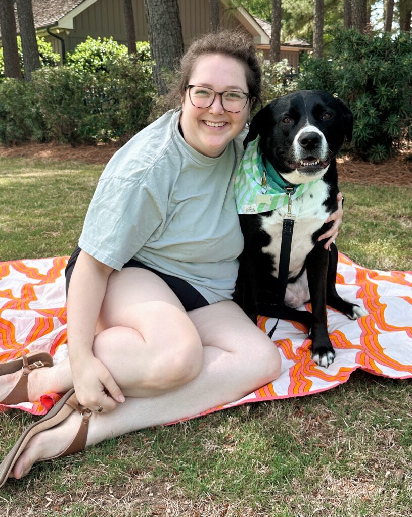 Susannah Brinkley Henry, wearing glasses, sits on a patterned blanket outdoors, smiling and posing with a black and white dog sporting a light green bandana.