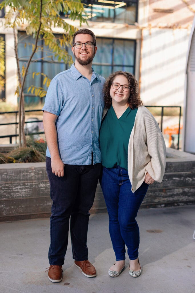 A man and Susannah Brinkley Henry stand side by side outdoors, smiling at the camera. They are casually dressed and positioned in front of a concrete planter and a modern building.